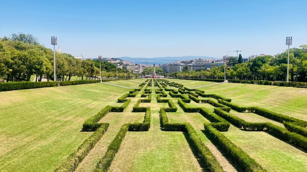 An intricate hedge maze in a lush green park in Lisbon, Portugal, with a cityscape view in the background on a clear spring day.