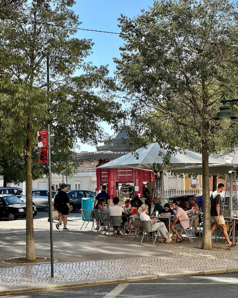 Lisbon in April: The Perfect Time To Visit 7 Vibrant outdoor café in Lisbon with people enjoying sunny weather, surrounded by trees and historic architecture, capturing the lively atmosphere of Lisbon in April.