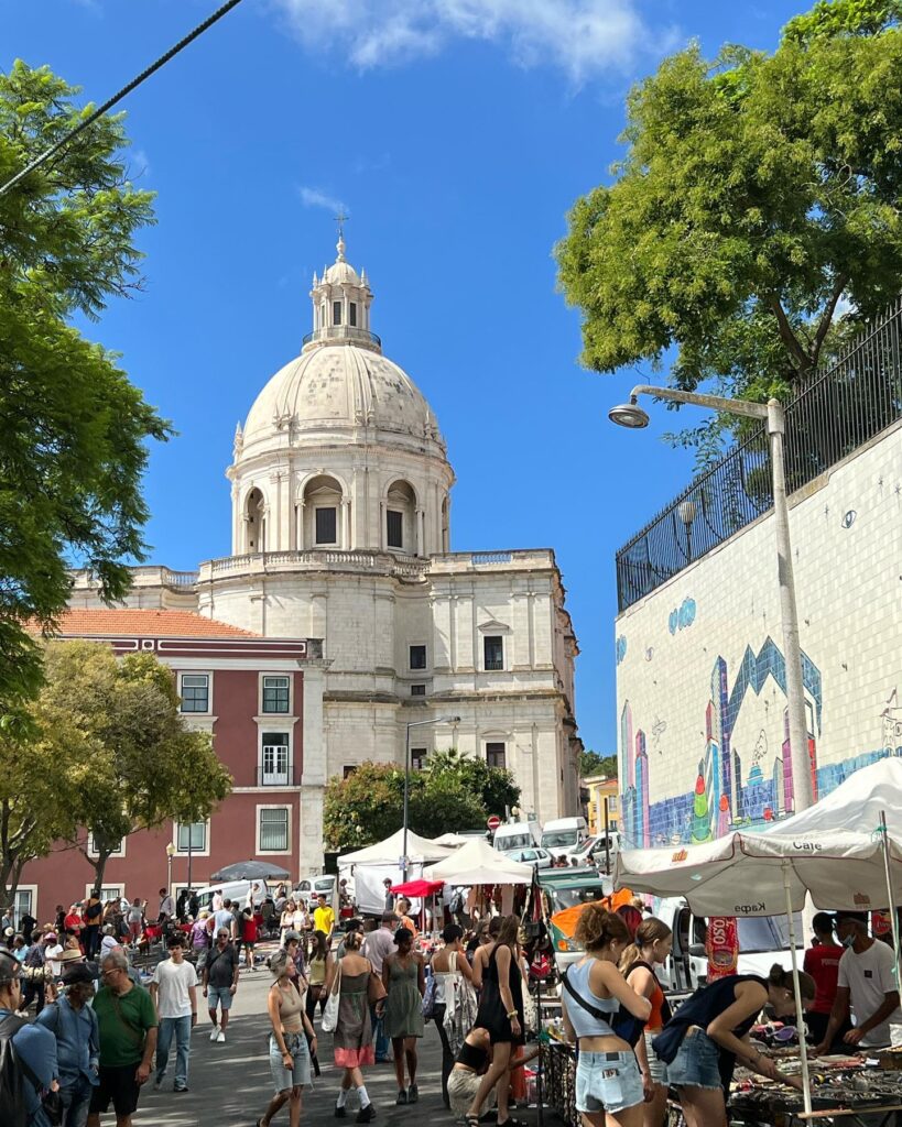 Alfama in Lisbon, Portugal, bustling with tourists exploring historic streets, local markets, and vibrant cafes under a clear blue sky.