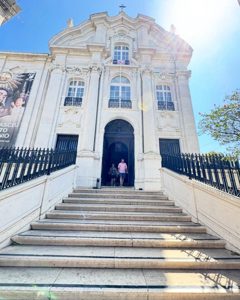 Alfama Church in Lisbon, Portugal, showcasing Baroque architecture with ornate details and a grand entrance, set against a bright blue sky on a sunny day.