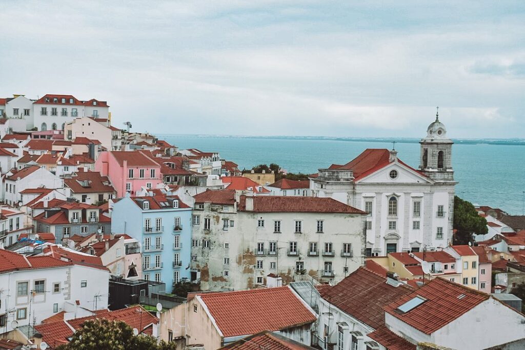 Alfama district in Lisbon with colorful historic buildings, narrow streets, and the Tagus River in the background, showcasing the charm and vibrant atmosphere of this iconic neighborhood.
