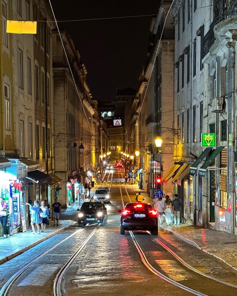 Night view of Alfama district in Lisbon with tram tracks, illuminated shops, and pedestrians enjoying the vibrant atmosphere.