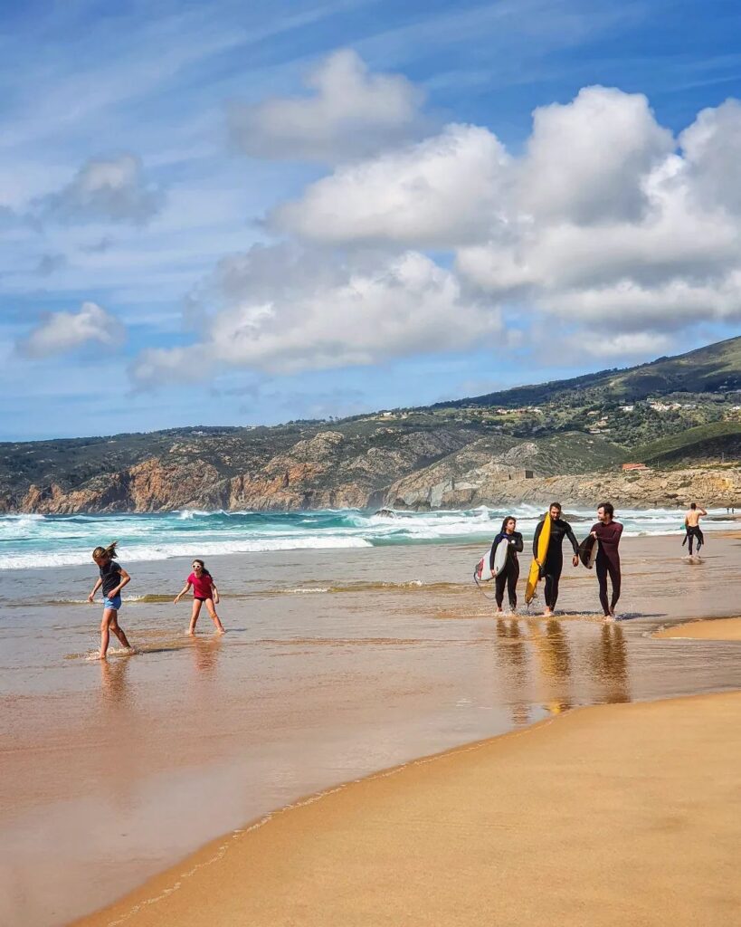 Scenic Lisbon beach scene featuring families and friends relaxing and walking along the sandy shore under a partly cloudy sky.