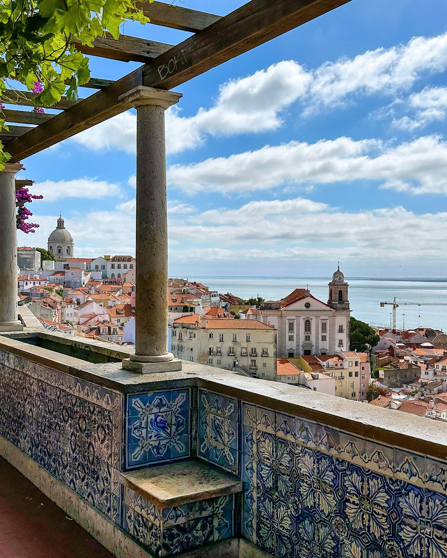 Scenic view of Alfama district in Lisbon from a balcony with traditional azulejos tiles, overlooking colorful rooftops and the Tagus River under a bright blue sky.