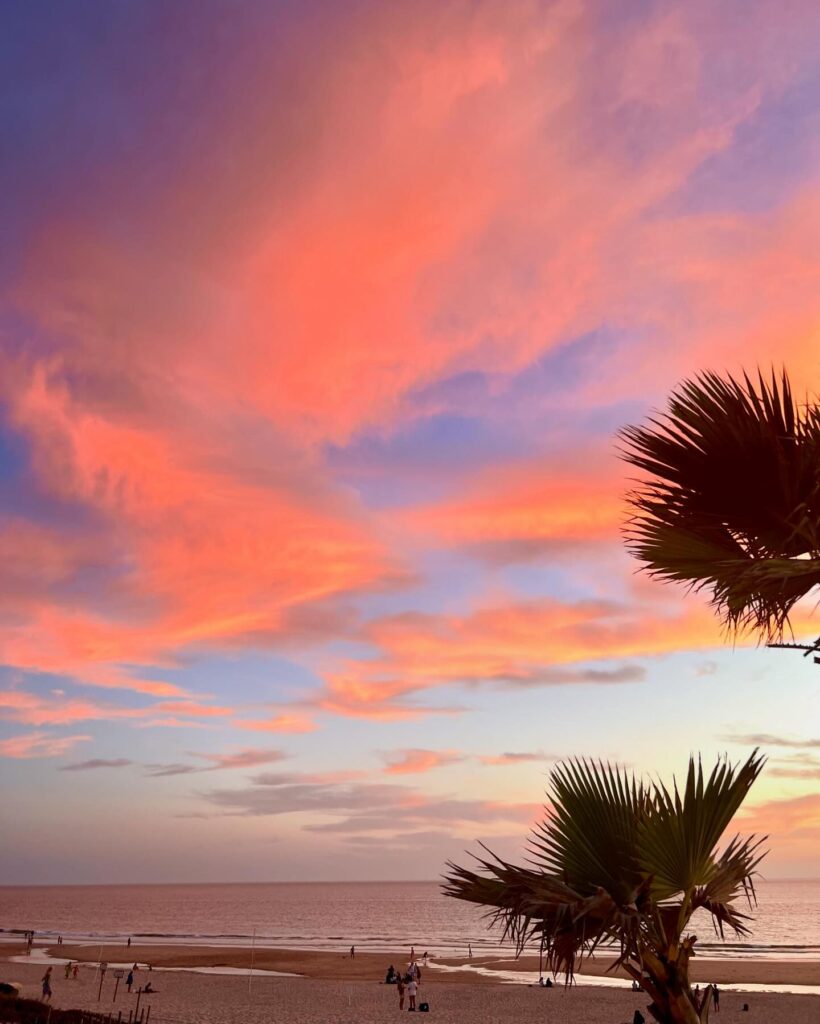 Sunset over Lisbon's beaches showcasing vibrant pink and orange clouds, palm trees, and calm ocean waters, perfect for relaxing seaside visits in Lisbon.