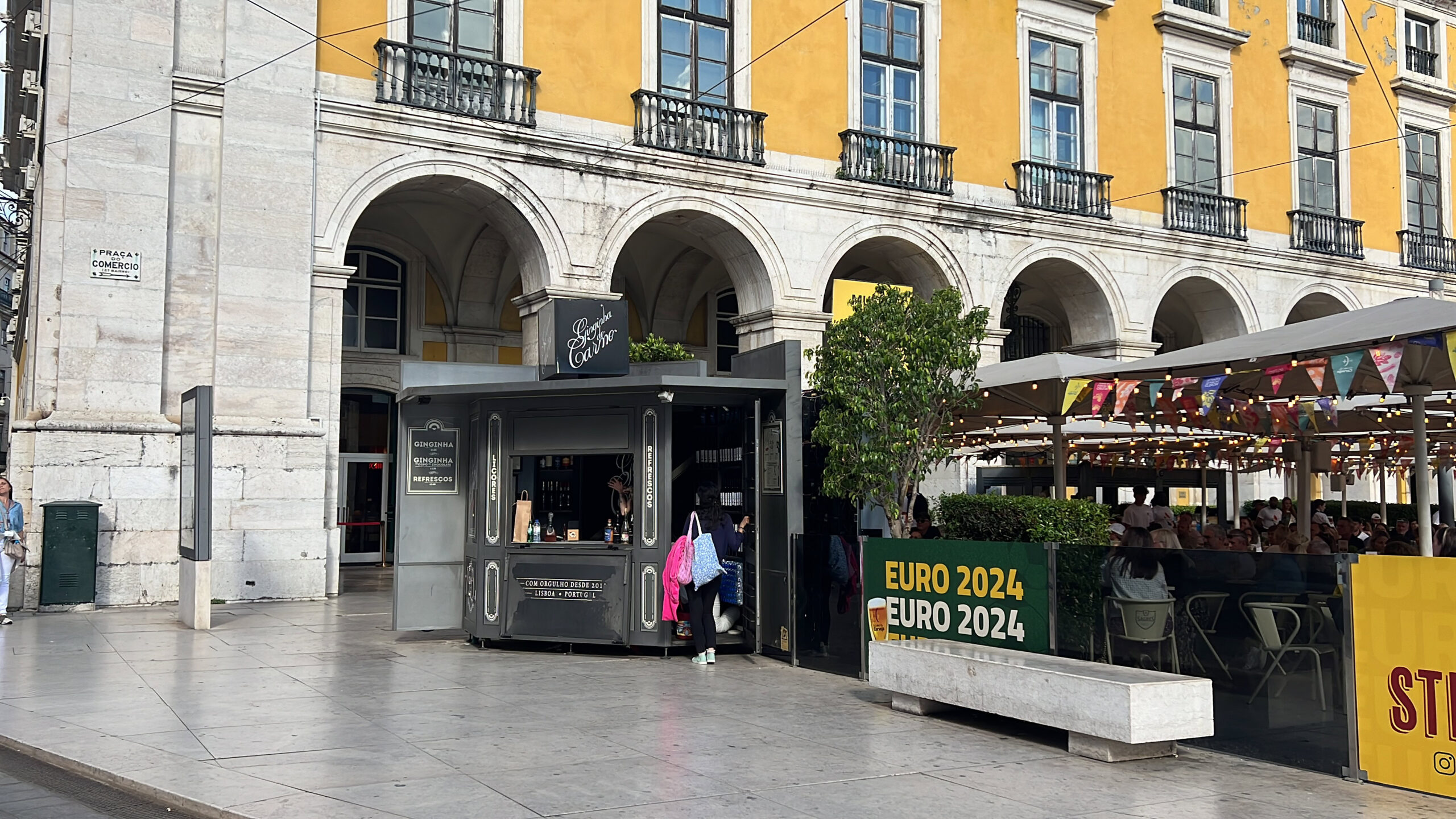 Ginjinha in Lisbon: 4 Authentic Places to Drink It 7 Historic Lisbon building with vibrant yellow facade, lively outdoor café area, and a city kiosk promoting EURO 2024, capturing the essence of LisboaVibes and Lisbon's bustling urban atmosphere.