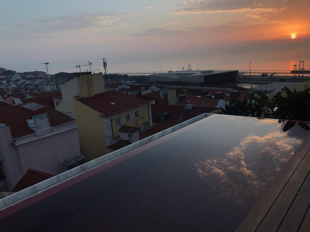 Scenic sunset view over Alfama district in Lisbon from a rooftop terrace, showcasing colorful rooftops, the Tagus River, and the iconic 25 de Abril Bridge at dusk.