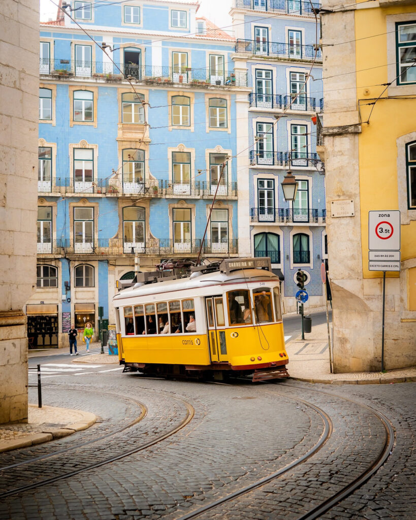 Vintage yellow tram navigating through Alfama district in Lisbon, Portugal, with vibrant pastel buildings and cobblestone streets.