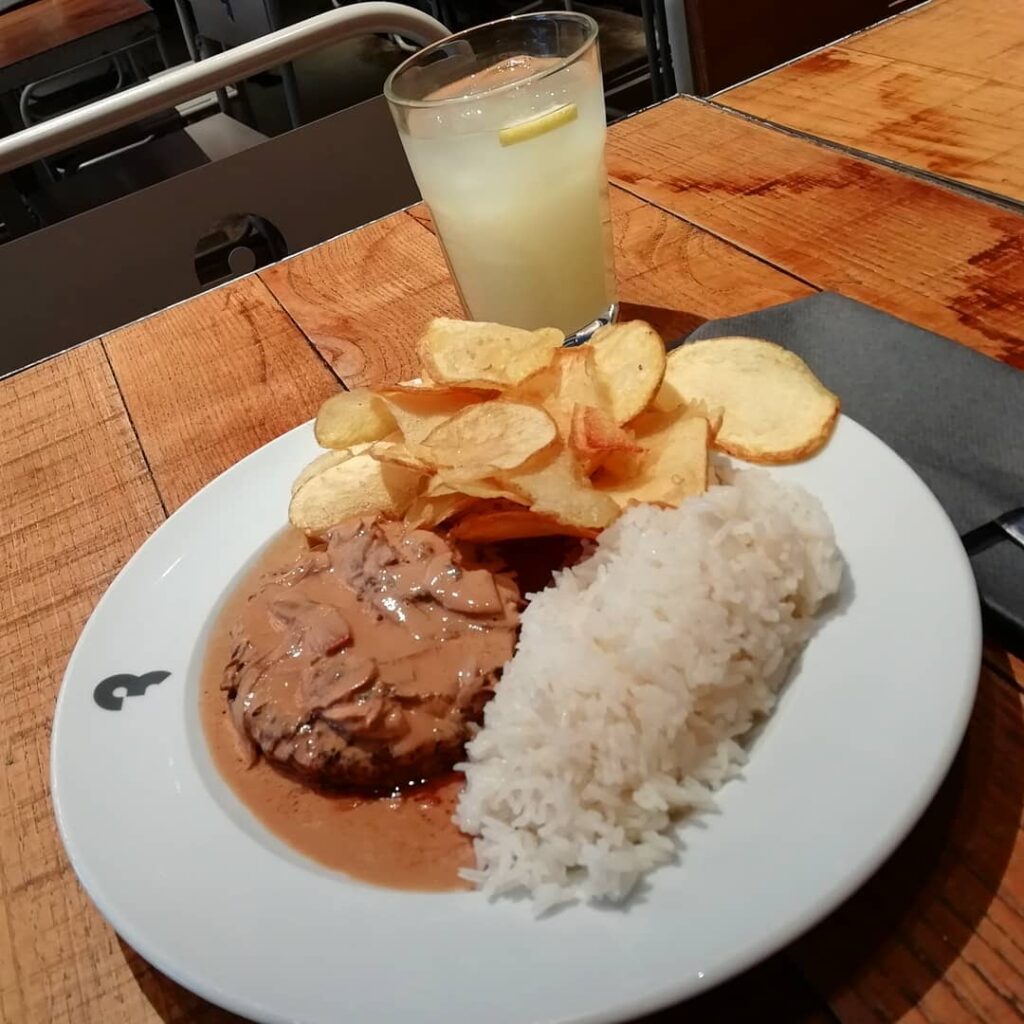 Creamy mushroom sauce over tender beef steak with white rice, crispy potato chips, and a refreshing lemonade with lemon slices on a wooden table in Lisbon. Perfect for a relaxing Lisbon dining experience.