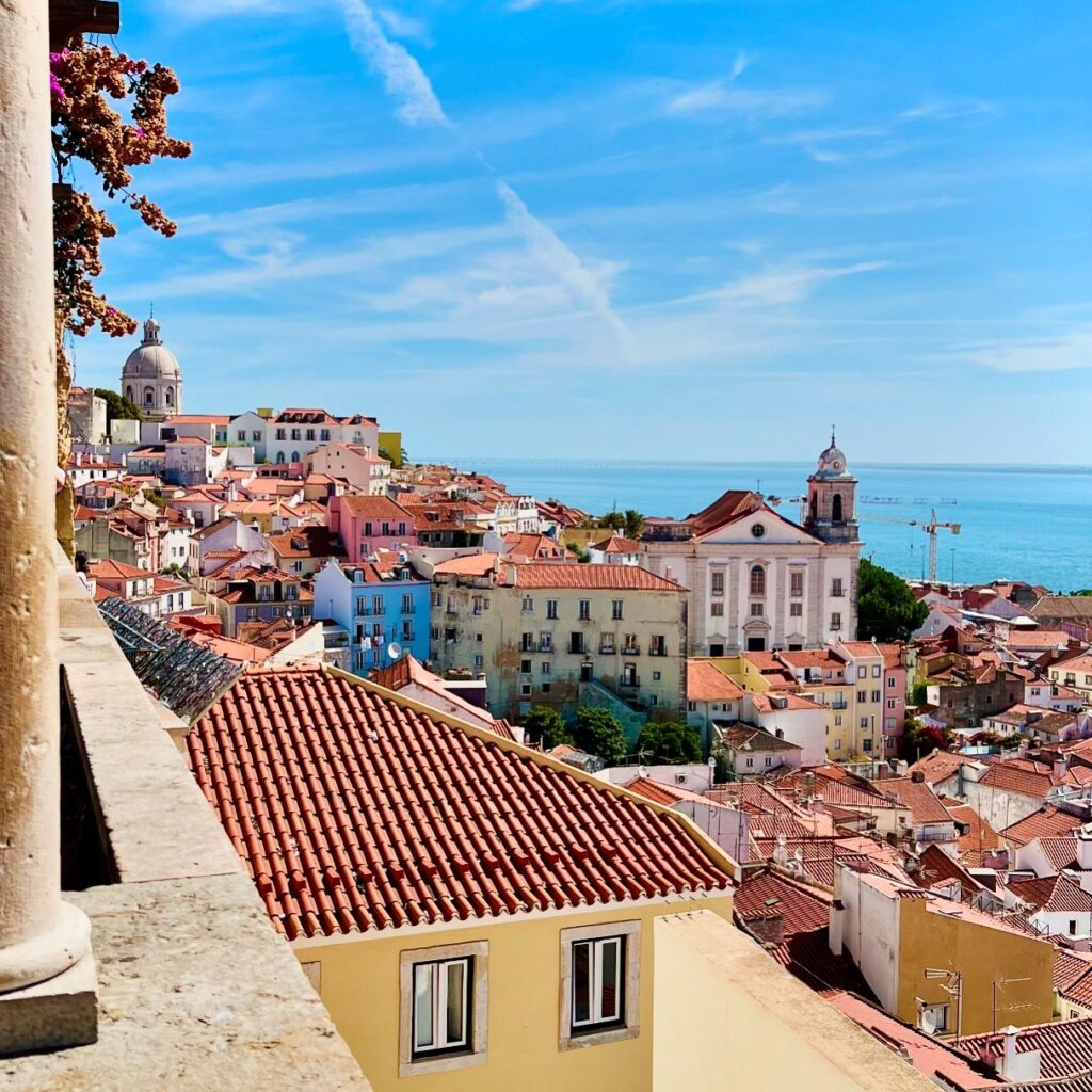 Panoramic view of Lisbon's colorful rooftops, historic buildings, and the Atlantic Ocean in the background, showcasing the city's vibrant architecture and scenic coastal setting.