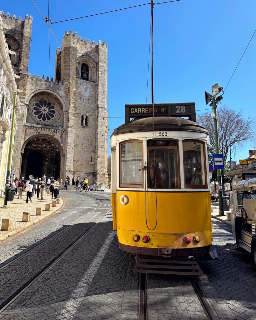Iconic yellow Tram 28 navigating through historic Lisbon streets near a medieval church, showcasing the classic Lisbon tram experience and vibrant city atmosphere.