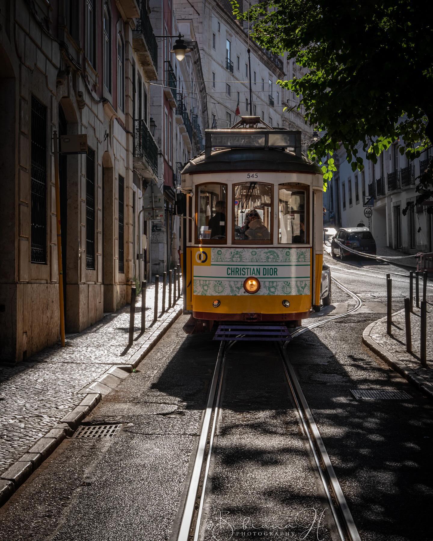 Vintage Lisbon tram navigating narrow city streets with traditional architecture and lush greenery.